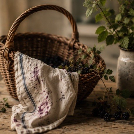 A wicker basket of foraged blackberries with a stained linen cloth on a wooden table