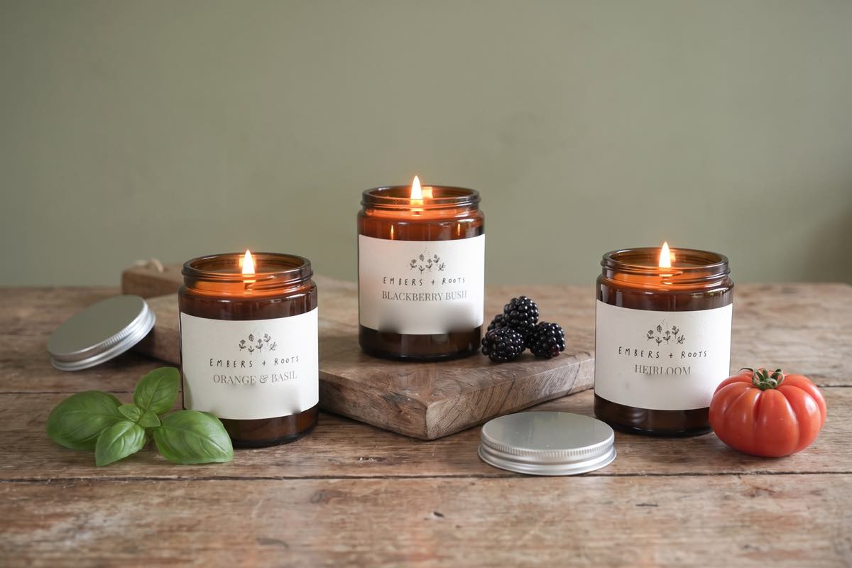 All three summer candles lit together on a wooden table: Orange & Basil beside a sprig of basil, Blackberry Bush with blackberries, Heirloom with a ripe tomato